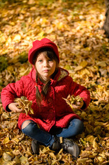 Beautiful little girl in red jacket is sitting on yellow leaves and playing on warm and sunny autumn day. Happy childhood concept