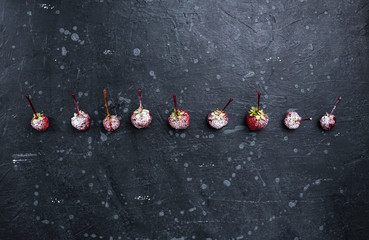 Berries of ripe strawberries in powdered sugar on sticks for snacks are laid out in a row on a dark stone surface. Top view.