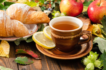 Mug of tea with lemon, croissant, autumn fall leaves, fruits on wooden background. Cozy home breakfast, autumn hygge style, comfort concept.