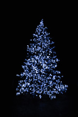 Silhouette of blurry Christmas tree illuminated and decorated with tiny blue fairy string lights in dark night creating beautiful bokeh effect with glowing out of focus circles and shiny dots