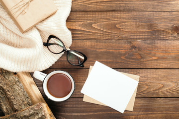 Flatlay composition with white knitted scarf, cup of tea, firewood, letter, blank paper card, glasses on wooden desk table. Hygge style, cozy autumn or winter concept. Flat lay, top view, overhead.