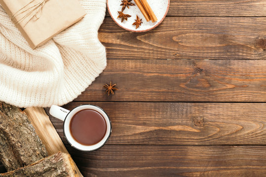 Flatlay composition with white knitted scarf, cup of tea, firewood, cinnamon sticks, gift box on wooden desk table. Hygge style, cozy autumn or winter holiday concept. Flat lay, top view, overhead.