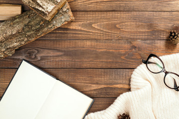 Feminine desk with white knitted scarf, glasses, firewood, book with empty pages on wooden background. Hygge, nordic, scandinavian style workplace.