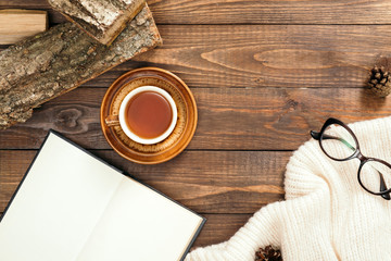 Hygge style flatlay composition with cup of tea, book with empty pages, white knitted scarf, woman glasses, firewood on wooden desk table. Autumn or winter holidays concept. Flat lay, top view.