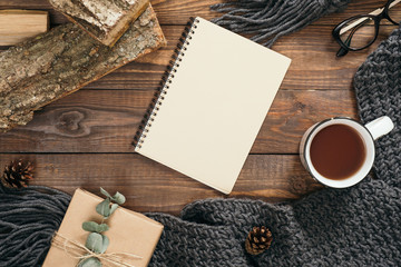 Flatlay hygge style composition with fashion women knitted scarf, blank paper notebook, mug of tea, gift box, firewood on wooden background. Flat lay, top view, overhead. Cozy autumn home desk