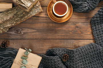 Hygge style flatlay frame with cup of tea, fashion knitted scarf, gift box, firewood on wooden background. Flat lay, top view, overhead. Cozy autumn home desk concept