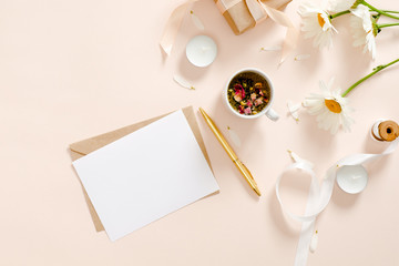 Romantic flatlay composition with blank paper card mockup, envelope, herbal tea, ribbon, daisy chamomile flower on pastel pink background. Feminine home office desk concept. Flat lay, top view.