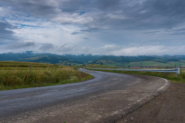 Empty, curved asphalt road after rain , stormclouds on the sky, village in the background in Transylvania, Romania.