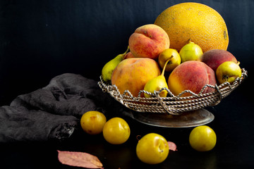 In the fruit bowl lie peaches, plums, pears, melons. Near dark fabric, black background. Still life.