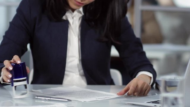 Tilting up shot of attentive mixed race lady, wearing jacket and white blouse, sitting at desk in notary office, checking signed contract copy and sealing it with self-inking rubber stamp