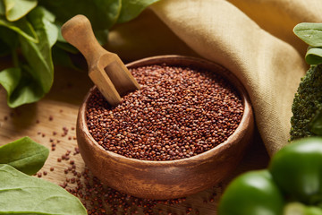 close up view of red quinoa seeds in wooden bowl with spatula near beige napkin and green vegetables