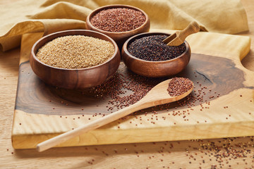 white, black and red quinoa in wooden bowls and spoon on wooden chopping board near napkin