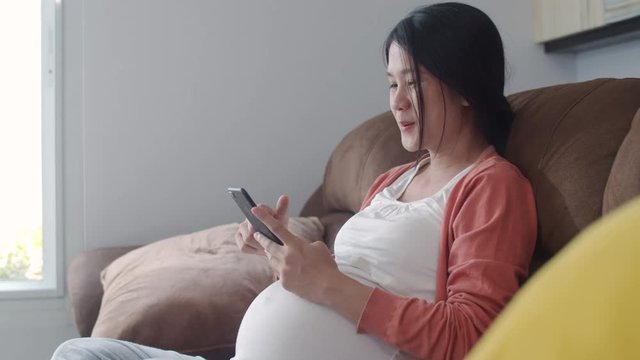Young Asian Pregnant woman using mobile phone search pregnancy information. Mom feeling happy smiling positive and peaceful while take care her child lying on sofa in living room at home concept.