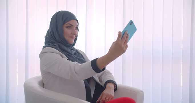 Closeup side view portrait of young muslim attractive female doctor in hijab and white coat taking selfies on the phone smiling happily sitting in the armchair in the white room indoors