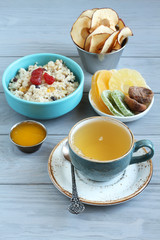 Oatmeal with fruit pieces and candied strawberries, pieces of candied pineapple, kiwi and mango, dried figs, honey and apple chips in small bowls and a cup of green tea on a gray wooden table. 