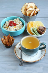 Oatmeal with fruit pieces and candied strawberries, pieces of candied pineapple, kiwi and mango, dried figs, apple and pear chips in small bowls and a cup of green tea on a gray wooden table. 