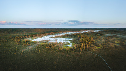  Warmly colored sunrise over a foggy swamp. Aerial view of stunning landscape at peat bog at Cenas Tirelis in Latvia. Wooden trail leading along the lake surrounded by pounds and forest. 