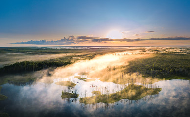 Obraz premium Warmly colored sunrise over a foggy swamp. Aerial view of stunning landscape at peat bog at Cenas Tirelis in Latvia. Wooden trail leading along the lake surrounded by pounds and forest. 