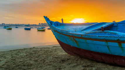 Obraz premium Turquoise blue fishing boat on the beach at sunrise with Alexandria skyline in far distance and colorful sky at sunrise