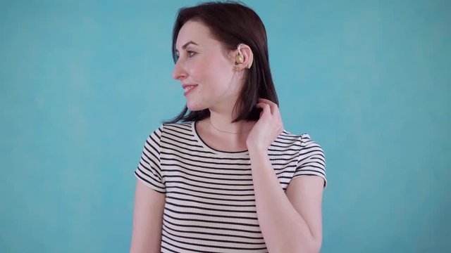 Smiling young woman with hearing impairment hears with hearing aid in ear on blue background