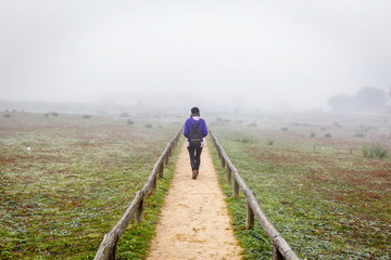 Fototapeta na wymiar Traveler woman walking alone on a field surrounded by fog. Alone woman from back walking through the fog in forest.