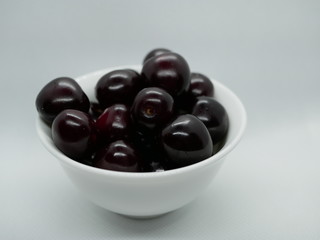 a handful of ripe maroon cherries in a small white porcelain bowl on a white background. cherry harvest. natural vitamin. vegetarian food. health food.