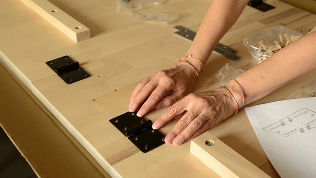 The hands of a Caucasian woman place a black metal hinge on two light wooden boards