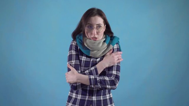 Frozen young woman in scarf and glasses can't get warm standing on blue background