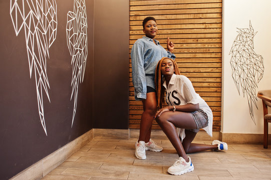 Two african womans in stylish casual clothes posing against wooden wall sat down and dance twerk.