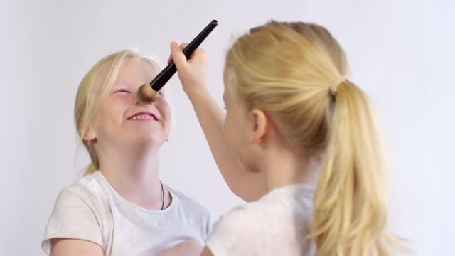 Chest-up shot of blonde Caucasian twin girls fooling around with make-up brush, with one pretending to powder and tickling her sister’s face, and another girl squirming and laughing
