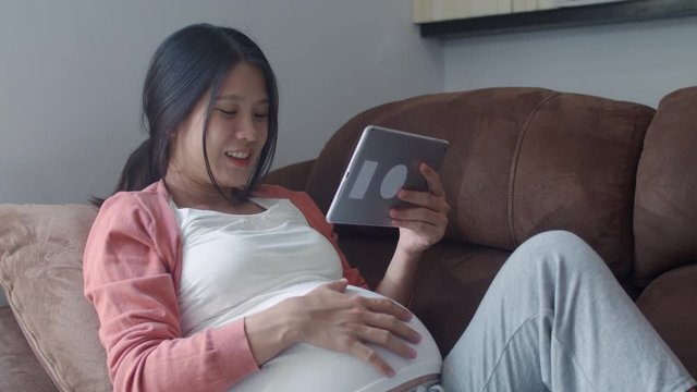 Young Asian Pregnant woman using tablet search pregnancy information. Mom feeling happy smiling positive and peaceful while take care her child lying on sofa in living room at home concept.