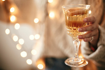  Stylish girl in cozy sweater holding champagne glass on background of christmas lights, celebrating and toasting in festive room. Space for text. Happy New Year eve party. Happy Holidays