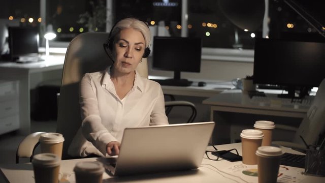 Waist-up shot of smiling grey-haired Caucasian businesswoman, dressed in white blouse and wearing headset, sitting alone late at night in dark office with coffee and participating in conference call