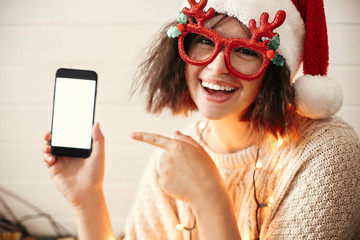 Stylish happy girl in festive glasses with reindeer horns smiling and showing phone empty screen in christmas lights. Young hipster woman in santa hat showing smartphone with space for text