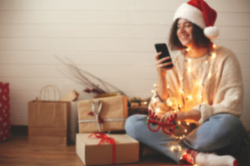 Blurred image of stylish happy girl in santa hat looking at phone screen in festive christmas lights on background o presents and gifts in modern room. Space for text. Happy Holidays