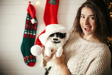 Stylish happy girl smiling and playing with cute cat in santa hat in background of  christmas tree lights and stockings. Young hipster woman in cozy sweater hugging kitty. Happy Holidays