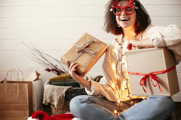 Stylish happy girl in festive glasses with reindeer horns holding christmas gift boxes and smiling...