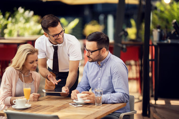 Smiling waiter showing the menu on touchpad to a couple in a cafe.