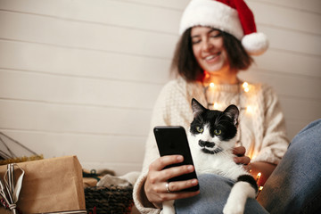 Stylish happy girl in santa hat with cute cat looking at phone screen on background of festive christmas lights and  presents. Young hipster woman hugging kitty with funny emotions