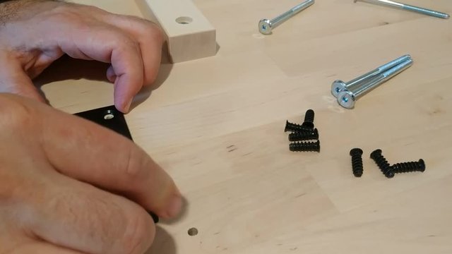 Fixing with a black metal hinge on two light wooden boards. One hand holds the hinge in place and the other positions the screws. 25Fps