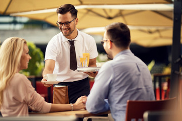 Happy waiter serving guests and giving them coffee in a cafe.