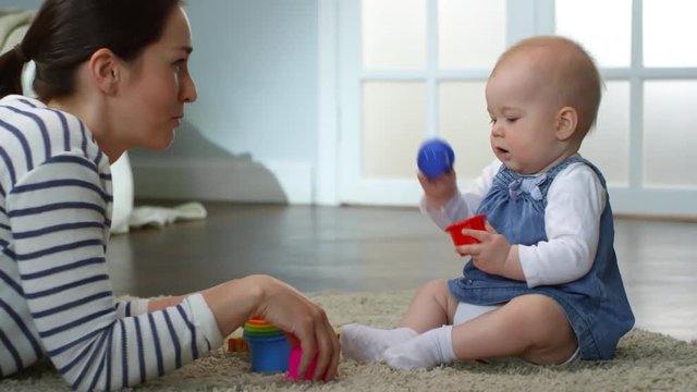 Full shot of adorable Caucasian baby girl sitting on beige carpet at home, playing with colorful stacking cups and being entertained by her mom, who is talking to baby and touching her nose