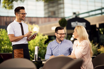 Happy waiter using touchpad while taking an order from a couple in a cafe.