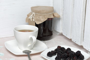 Jars of jam, covered with paper and tied with twine. On the string hang craft labels. Next to it is a plate of fresh blackberries and a cup of tea.