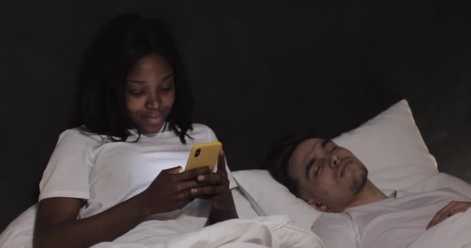 Married multi-ethnic couple lying in bed at night. Woman using smartphone texting with lover, while his husband is sleeping.
