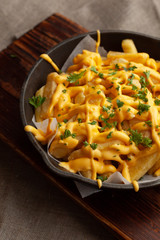 Mouthwatering hot french fries with a lot of cheese sauce with herbs in a pan, on a wooden board