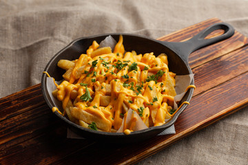 Mouthwatering hot french fries with a lot of cheese sauce with herbs in a pan, on a wooden board - obrazy, fototapety, plakaty