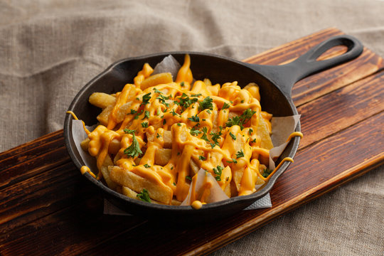 Mouthwatering hot french fries with a lot of cheese sauce with herbs in a pan, on a wooden board