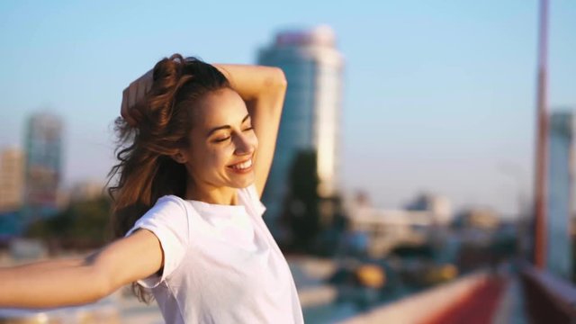 happy smiling young pretty woman in white t-shirt is standing on the bridge on blue sky background, hand straightens hair, enjoying peaceful sunset and looking at camera. calm and relaxation. slow