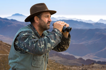 A hunter in a hat with binoculars looks out for prey.